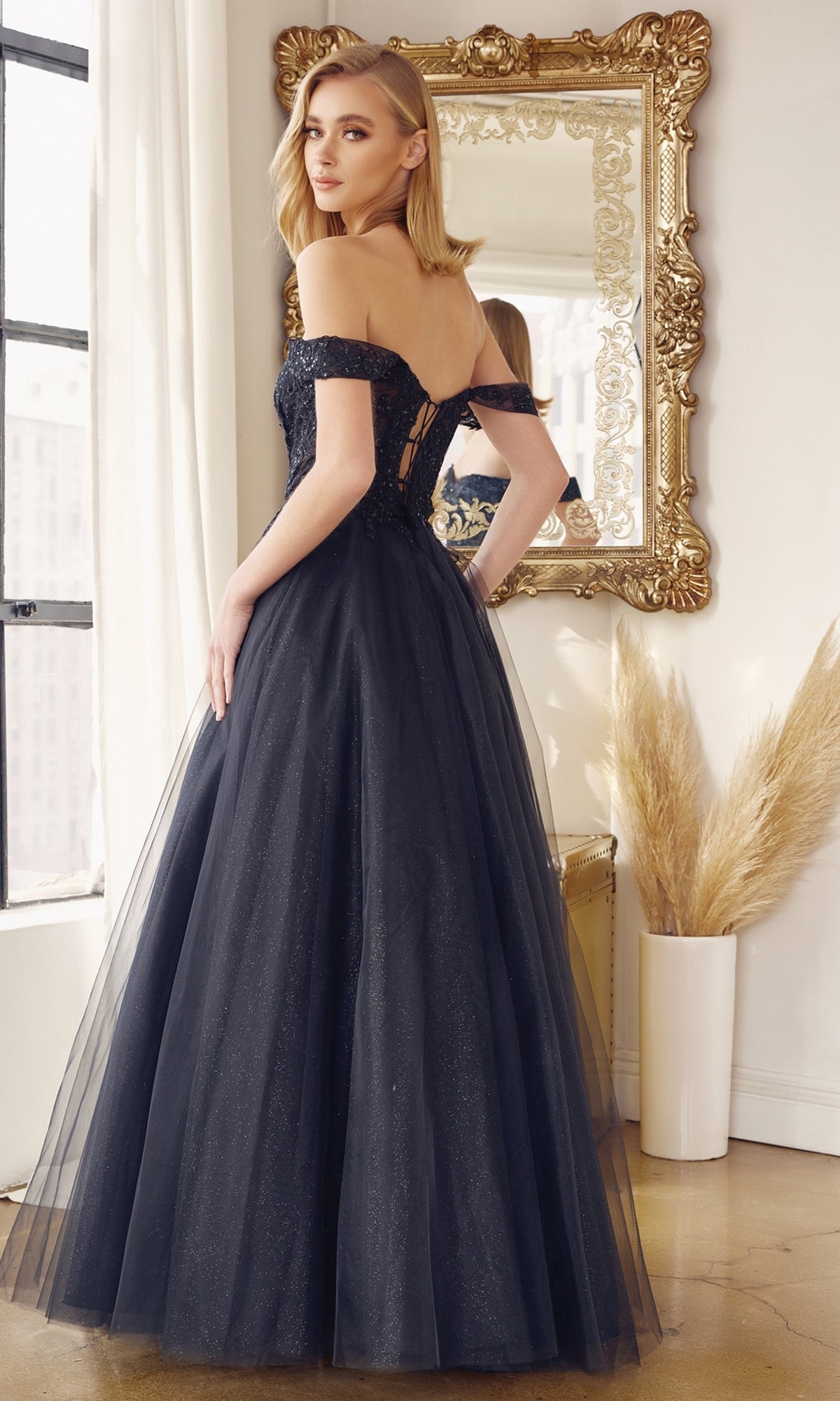 A woman in the Lace-Up Off-Shoulder Long A-Line Prom Dress 280 stands before a large ornate mirror in a bright room with tall windows and a vase of pampas grass.